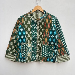 Puede incluir: Chaqueta acolchada con diseño patchwork en tonos verdes, turquesa y rojo. La chaqueta presenta cuello mandarín, dos bolsillos delanteros y forro a rayas. La chaqueta está colgada de una percha de madera.