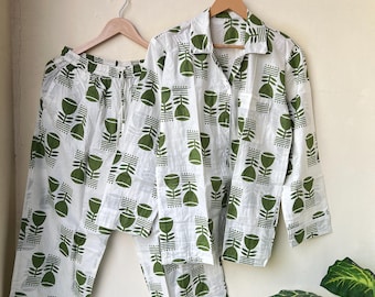 May include: White pajama set with a green floral pattern. The set includes a button-down shirt with a collar and long sleeves, and matching pants with a drawstring waist. The pajamas are hanging on wooden hangers.