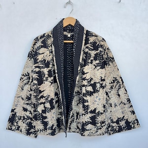 Può includere: Giacca fantasia nera e beige con colletto e fodera neri a contrasto. La giacca presenta un motivo floreale ed è appesa a una gruccia di legno contro un muro bianco.