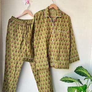 May include: A two-piece pajama set in a light green color with a repeating floral pattern in pink and brown. The long-sleeved top has white piping and buttons. The pants have a drawstring waist and white piping.
