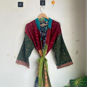 Pode incluir: Um roupão tipo quimono de patchwork com uma mistura de tecidos em vermelho escuro, verde escuro e azul com padrões florais e geométricos. A peça tem mangas largas, uma gola em tom azul-petróleo e uma faixa de tecido verde-claro na cintura.