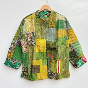 Op de afbeelding: Een groen, geel en bruin patchwork gequilte jas met een knoopsluiting. De jas heeft een bloemenprint op de mouwen en een zak op de voorkant.