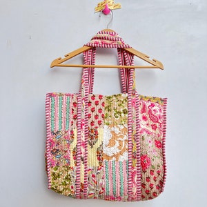 May include: A patchwork tote bag with pink, green and white floral patterns. The bag has two long straps and is hanging on a wooden hanger.