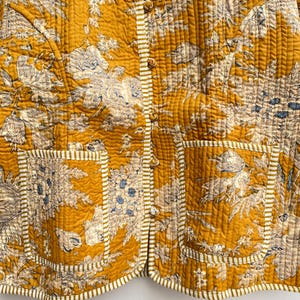 May include: A mustard yellow quilted jacket with a floral print and white trim. The jacket has a mandarin collar, a button closure, and two patch pockets.