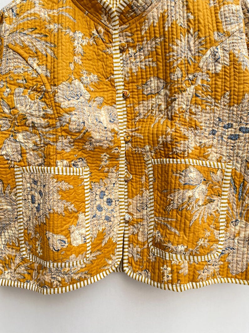 May include: A mustard yellow quilted jacket with a floral print and white trim. The jacket has a mandarin collar, a button closure, and two patch pockets.