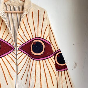 Puede incluir: Chaqueta color crema con cremallera completa y dise&ntilde;os de ojos bordados. Los ojos son de color rojo oscuro con pupilas negras y contornos rosas, rodeados de l&iacute;neas marrones radiantes. La chaqueta tiene un forro a rayas y est&aacute; colgada de una percha de madera.