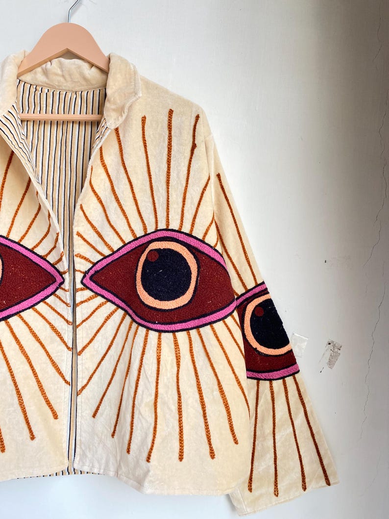 Puede incluir: Chaqueta color crema con cremallera completa y dise&ntilde;os de ojos bordados. Los ojos son de color rojo oscuro con pupilas negras y contornos rosas, rodeados de l&iacute;neas marrones radiantes. La chaqueta tiene un forro a rayas y est&aacute; colgada de una percha de madera.