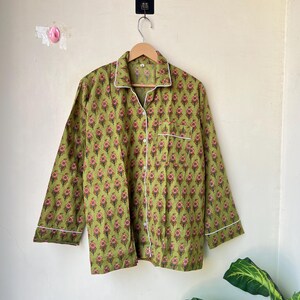 May include: Green floral print pajama top with white trim and buttons. The long-sleeved shirt is hanging on a wooden hanger. The floral pattern features pink and red flowers on a green background.