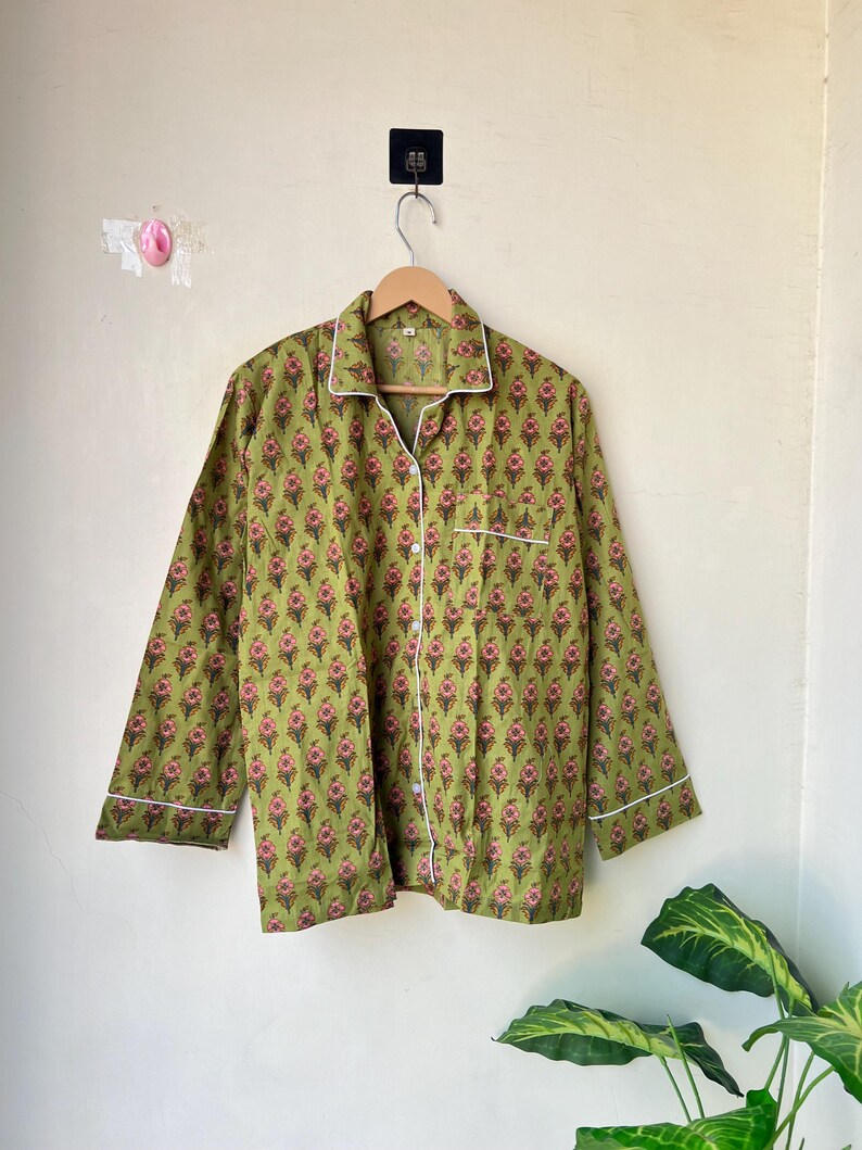 May include: Green floral print pajama top with white trim and buttons. The long-sleeved shirt is hanging on a wooden hanger. The floral pattern features pink and red flowers on a green background.