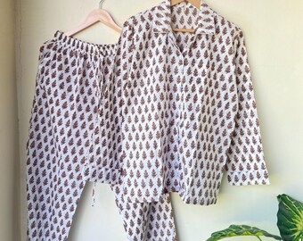 Op de afbeelding: Een witte pyjamaset met een bruin bladerpatroon, bestaande uit een overhemd met knopen en een broek met trekkoord, hangt aan een houten hanger. De set wordt gepresenteerd tegen een lichtgekleurde muur, met een plant in de hoek.