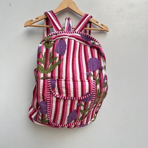 Puede incluir: Una mochila de rayas rosa y blancas con flores moradas bordadas. La mochila tiene un ribete blanco y una cremallera.
