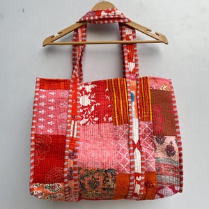 May include: A large patchwork tote bag with a red, pink, and orange color scheme. The bag is made of quilted fabric with various floral and geometric patterns. The bag has two long straps and is hanging on a wooden hanger.