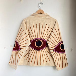 Puede incluir: Chaqueta beige con dise&ntilde;os de ojos bordados en granate, rosa y marr&oacute;n. La chaqueta tiene cuello y mangas largas. Los dise&ntilde;os de ojos est&aacute;n en la espalda y las mangas, con l&iacute;neas radiantes. La chaqueta est&aacute; colgada en una percha de madera.