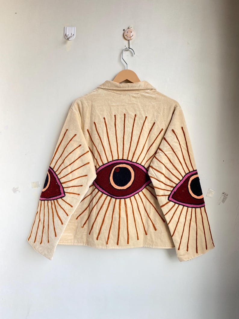 Puede incluir: Chaqueta beige con dise&ntilde;os de ojos bordados en granate, rosa y marr&oacute;n. La chaqueta tiene cuello y mangas largas. Los dise&ntilde;os de ojos est&aacute;n en la espalda y las mangas, con l&iacute;neas radiantes. La chaqueta est&aacute; colgada en una percha de madera.