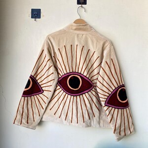 Puede incluir: Chaqueta beige con dise&ntilde;os de ojos bordados en burdeos, negro y marr&oacute;n. La chaqueta tiene cuello alto y mangas largas, con motivos de ojos en la espalda y las mangas. La chaqueta est&aacute; colgada en una percha de madera.