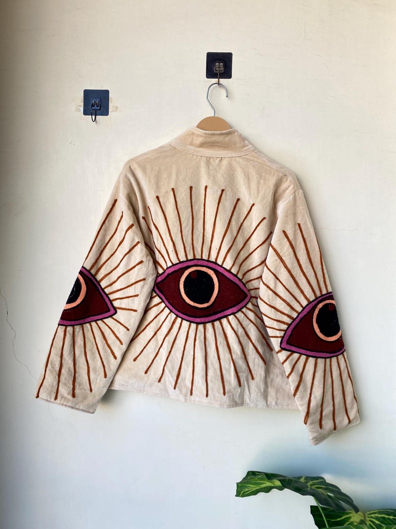 Puede incluir: Chaqueta beige con dise&ntilde;os de ojos bordados en burdeos, negro y marr&oacute;n. La chaqueta tiene cuello alto y mangas largas, con motivos de ojos en la espalda y las mangas. La chaqueta est&aacute; colgada en una percha de madera.