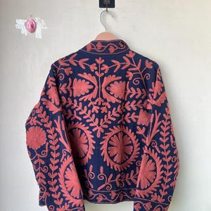 May include: A navy blue jacket with intricate coral-colored floral embroidery. The jacket is hanging on a wooden hanger against a plain white wall. The embroidery features a detailed pattern of flowers and leaves.