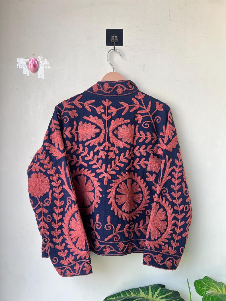 May include: A navy blue jacket with intricate coral-colored floral embroidery. The jacket is hanging on a wooden hanger against a plain white wall. The embroidery features a detailed pattern of flowers and leaves.