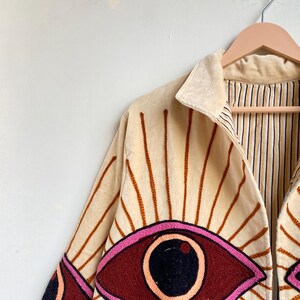 Puede incluir: Chaqueta color crema con dise&ntilde;o de ojo bordado. La chaqueta presenta un cuello marr&oacute;n claro y l&iacute;neas verticales que irradian del dise&ntilde;o del ojo. El ojo est&aacute; delineado en rosa con un iris rojo oscuro y una pupila negra. La chaqueta est&aacute; colgada en una percha de madera.
