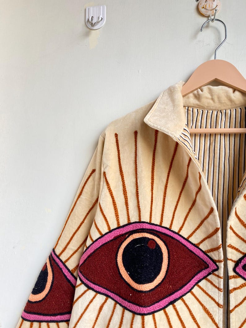 Puede incluir: Chaqueta color crema con dise&ntilde;o de ojo bordado. La chaqueta presenta un cuello marr&oacute;n claro y l&iacute;neas verticales que irradian del dise&ntilde;o del ojo. El ojo est&aacute; delineado en rosa con un iris rojo oscuro y una pupila negra. La chaqueta est&aacute; colgada en una percha de madera.