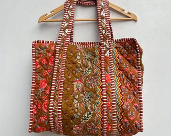 Op de afbeelding: Een bruine quilted tote bag met bloemen- en geometrische patronen. De tas heeft rode, witte en groene bies en twee lange hengsels.