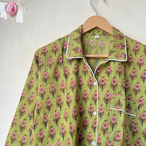 May include: Green pajama top with a floral pattern in pink and orange. The long-sleeved top has white trim on the collar, cuffs, and pocket. It is buttoned up and hanging on a wooden hanger.