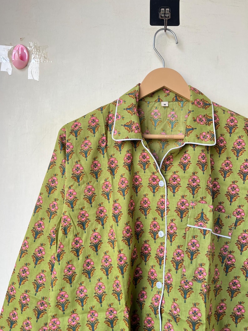 May include: Green pajama top with a floral pattern in pink and orange. The long-sleeved top has white trim on the collar, cuffs, and pocket. It is buttoned up and hanging on a wooden hanger.