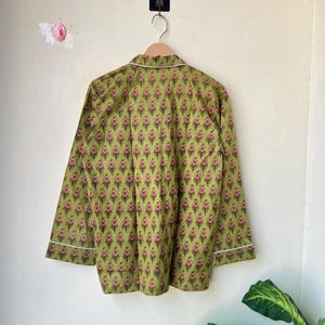 May include: A long-sleeved pajama top in a green floral pattern, hanging on a wooden hanger. The fabric features a repeating design of pink and brown floral motifs. White piping trims the collar and cuffs.