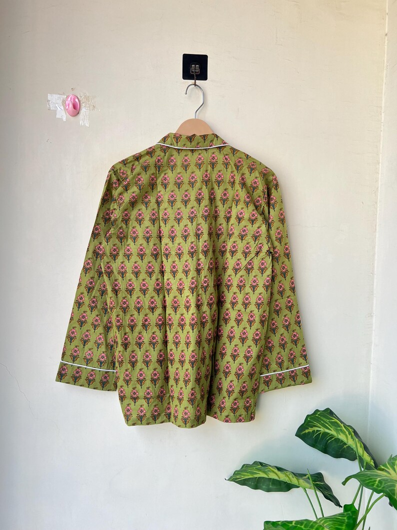 May include: A long-sleeved pajama top in a green floral pattern, hanging on a wooden hanger. The fabric features a repeating design of pink and brown floral motifs. White piping trims the collar and cuffs.