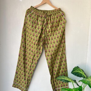 May include: Green pajama pants with a floral pattern in pink and brown. The pants have a drawstring waist and white piping along the edges. The pants are hanging on a wooden hanger.