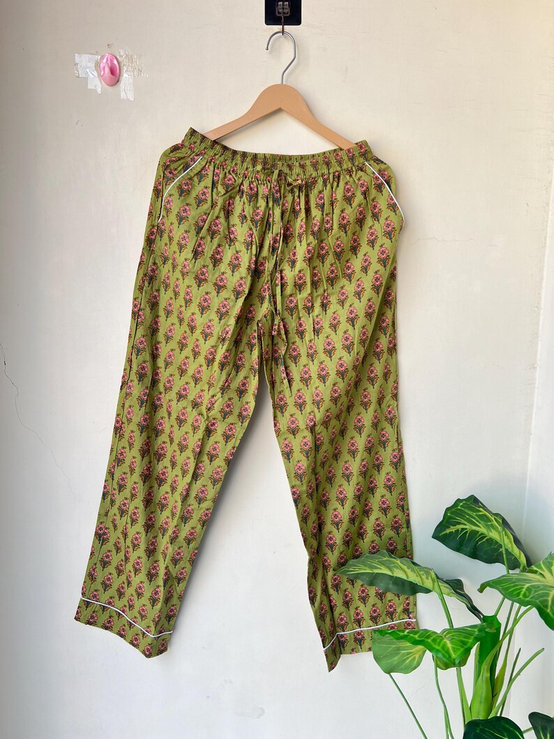 May include: Green pajama pants with a floral pattern in pink and brown. The pants have a drawstring waist and white piping along the edges. The pants are hanging on a wooden hanger.