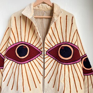 Puede incluir: Chaqueta color crema con dise&ntilde;os de ojos bordados. La chaqueta presenta grandes ojos estilizados en tonos burdeos, rosa y negro, con l&iacute;neas radiantes. Tiene una cremallera y un forro a rayas.