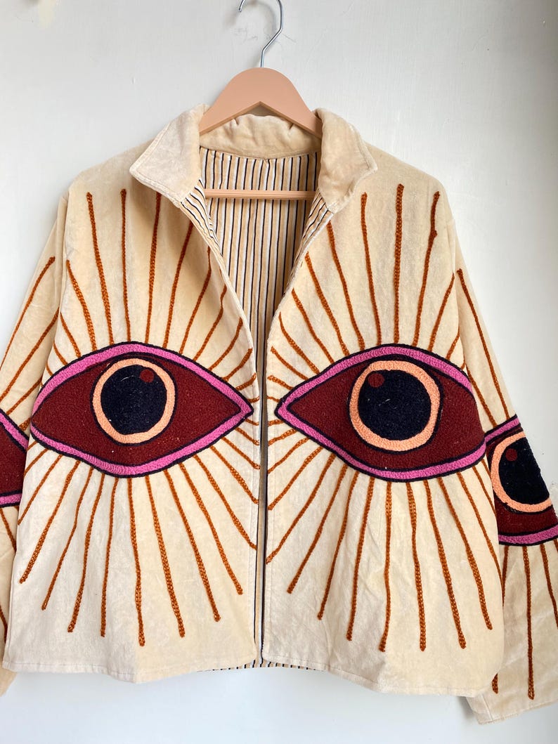 Puede incluir: Chaqueta color crema con dise&ntilde;os de ojos bordados. La chaqueta presenta grandes ojos estilizados en tonos burdeos, rosa y negro, con l&iacute;neas radiantes. Tiene una cremallera y un forro a rayas.