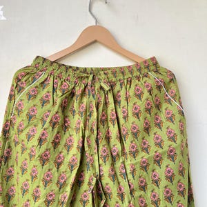 May include: Green floral print pajama pants with a drawstring waist and white trim. The pants are hanging on a wooden hanger against a neutral background. The floral pattern features pink flowers and green leaves.