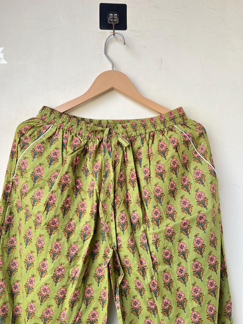 May include: Green floral print pajama pants with a drawstring waist and white trim. The pants are hanging on a wooden hanger against a neutral background. The floral pattern features pink flowers and green leaves.