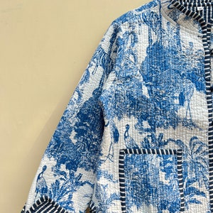 May include: A blue and white quilted jacket with a floral and bird print. The jacket has a mandarin collar, a front button closure, and a patch pocket on the left side. The jacket is trimmed with a black and white striped fabric.