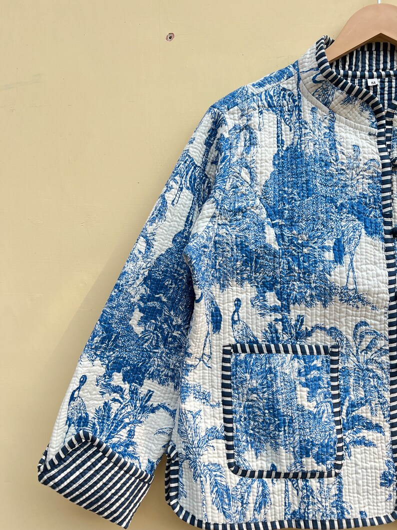 May include: A blue and white quilted jacket with a floral and bird print. The jacket has a mandarin collar, a front button closure, and a patch pocket on the left side. The jacket is trimmed with a black and white striped fabric.