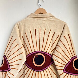 Puede incluir: Chaqueta de pana beige con dise&ntilde;os de ojos bordados en la espalda y las mangas. Los ojos est&aacute;n delineados en rosa y marr&oacute;n, con una pupila negra. La chaqueta tiene cuello y cierre de botones.