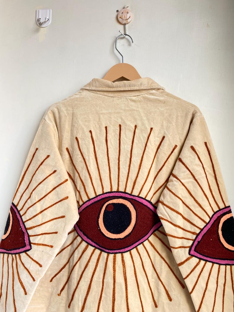 Puede incluir: Chaqueta de pana beige con dise&ntilde;os de ojos bordados en la espalda y las mangas. Los ojos est&aacute;n delineados en rosa y marr&oacute;n, con una pupila negra. La chaqueta tiene cuello y cierre de botones.