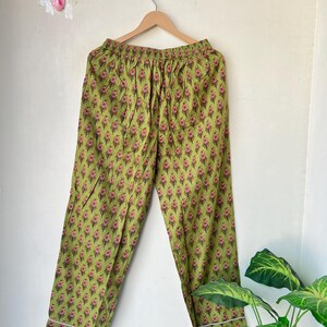 May include: Green pajama pants with a floral pattern in pink and brown. The pants have a white trim along the cuffs and are hanging on a wooden hanger. The background is a plain white wall.