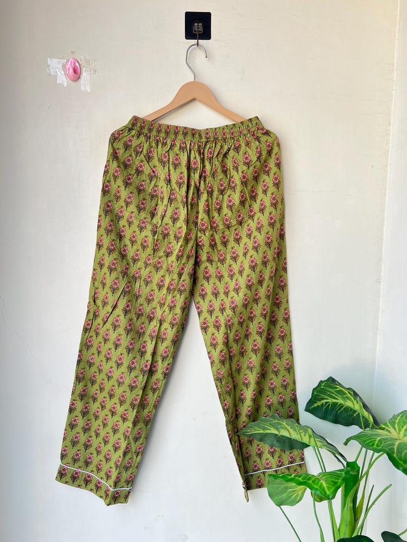 May include: Green pajama pants with a floral pattern in pink and brown. The pants have a white trim along the cuffs and are hanging on a wooden hanger. The background is a plain white wall.