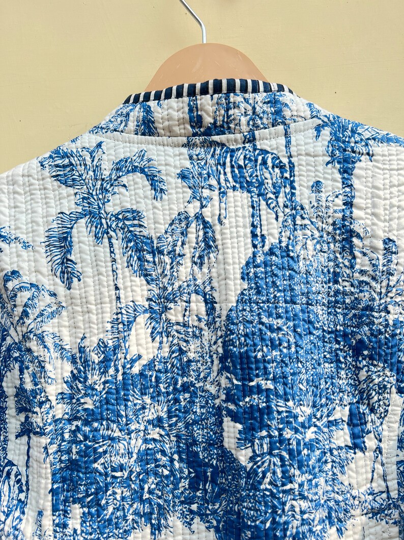 May include: A white quilted jacket with a blue and white tropical print featuring palm trees and animals. The jacket has a black and white striped trim around the collar.