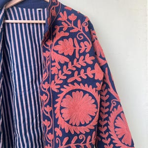 May include: A navy blue jacket with coral floral embroidery. The jacket features a striped lining in white and red. The embroidery is detailed and covers the front and sleeves of the jacket. The jacket is hanging on a wooden hanger.