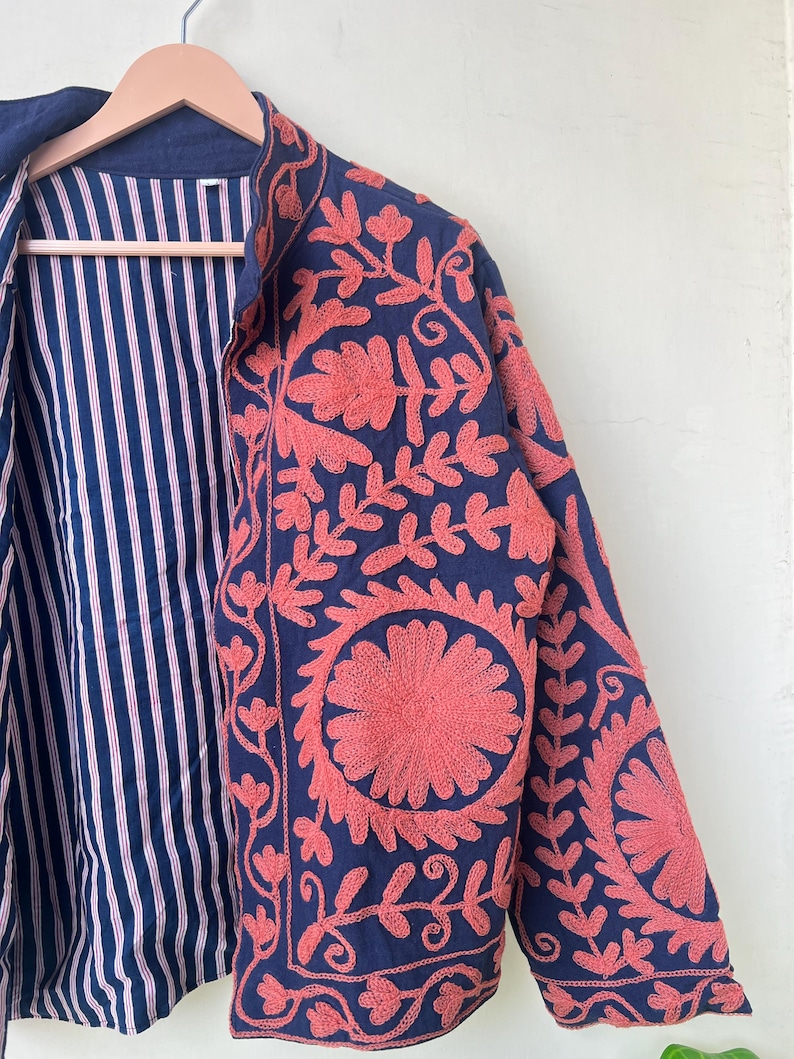 May include: A navy blue jacket with coral floral embroidery. The jacket features a striped lining in white and red. The embroidery is detailed and covers the front and sleeves of the jacket. The jacket is hanging on a wooden hanger.