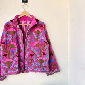 以下が含まれることがあります： 茶色、青、ピンクの色合いで、複雑な花の刺繍が施された鮮やかなピンク色のジャケット。 ジャケットはハイカラーで、木製のハンガーに吊るされています。 内側の裏地はストライプ柄です。