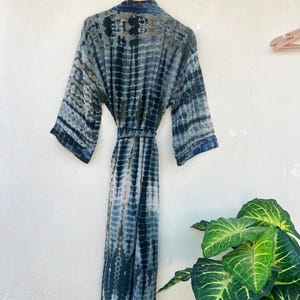 K&ouml;nnte beinhalten: Eine lange, blaue und wei&szlig;e Batik-Robe mit einem G&uuml;rtel. Die Robe h&auml;ngt an einem Kleiderb&uuml;gel an einer wei&szlig;en Wand.