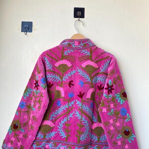 以下が含まれることがあります： マゼンタ色のジャケットで、複雑な花の刺繍が施されています。 ジャケットはハイカラーと長袖で、青、茶色、ピンクの色合いで詳細な刺繍が施されています。 ジャケットは木製のハンガーに吊るされています。