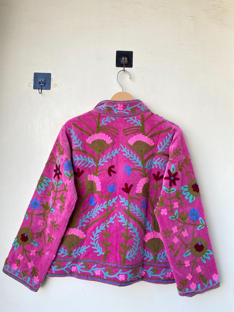以下が含まれることがあります： マゼンタ色のジャケットで、複雑な花の刺繍が施されています。 ジャケットはハイカラーと長袖で、青、茶色、ピンクの色合いで詳細な刺繍が施されています。 ジャケットは木製のハンガーに吊るされています。