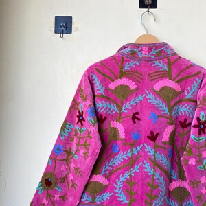 以下が含まれることがあります： ピンク色の鮮やかなジャケット。ピンク、青、茶色の花柄の刺繍が施されています。木製のハンガーに掛けられており、白い壁を背景にしています。刺繍は、花と葉の細かい模様が特徴です。
