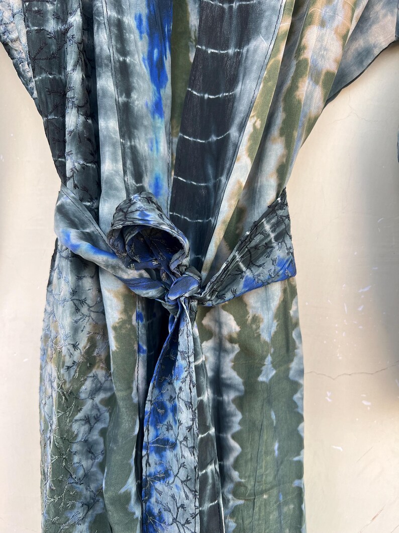 K&ouml;nnte beinhalten: Eine blaue und gr&uuml;ne Batik-Robe mit einem langen G&uuml;rtel, der in der Taille gebunden ist. Die Robe hat ein dezentes Stickmuster.